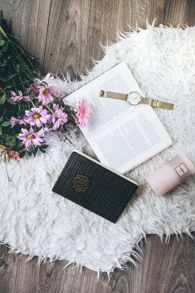 A cozy flatlay featuring a watch, open book, pink flowers, and faux fur rug.