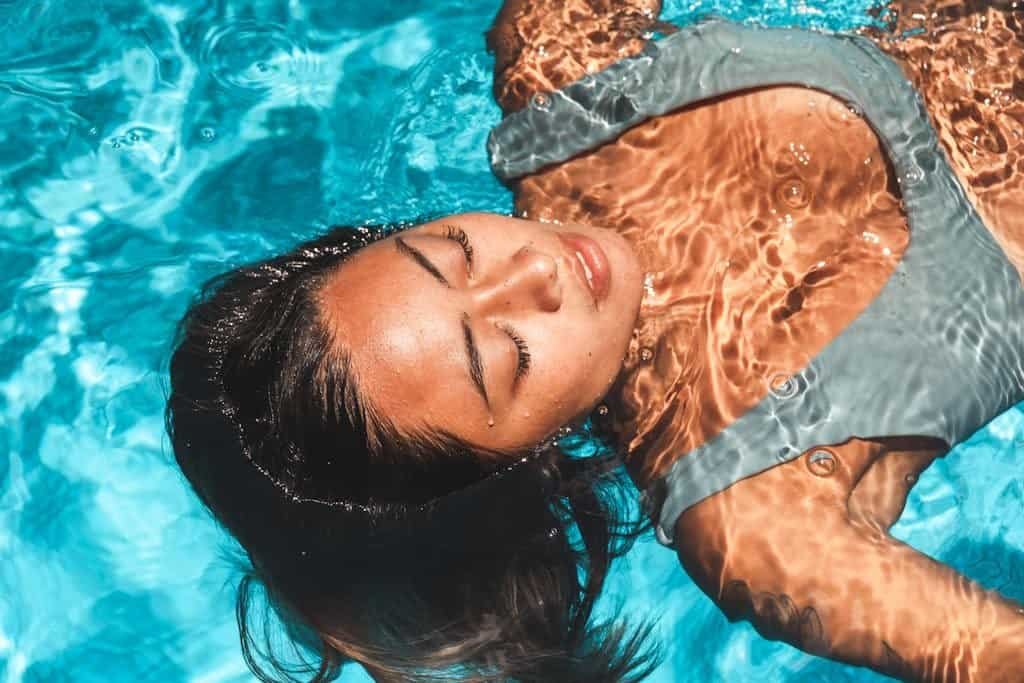 A serene portrait of an Asian woman floating with eyes closed in a vibrant blue pool, exuding relaxation and tranquility.